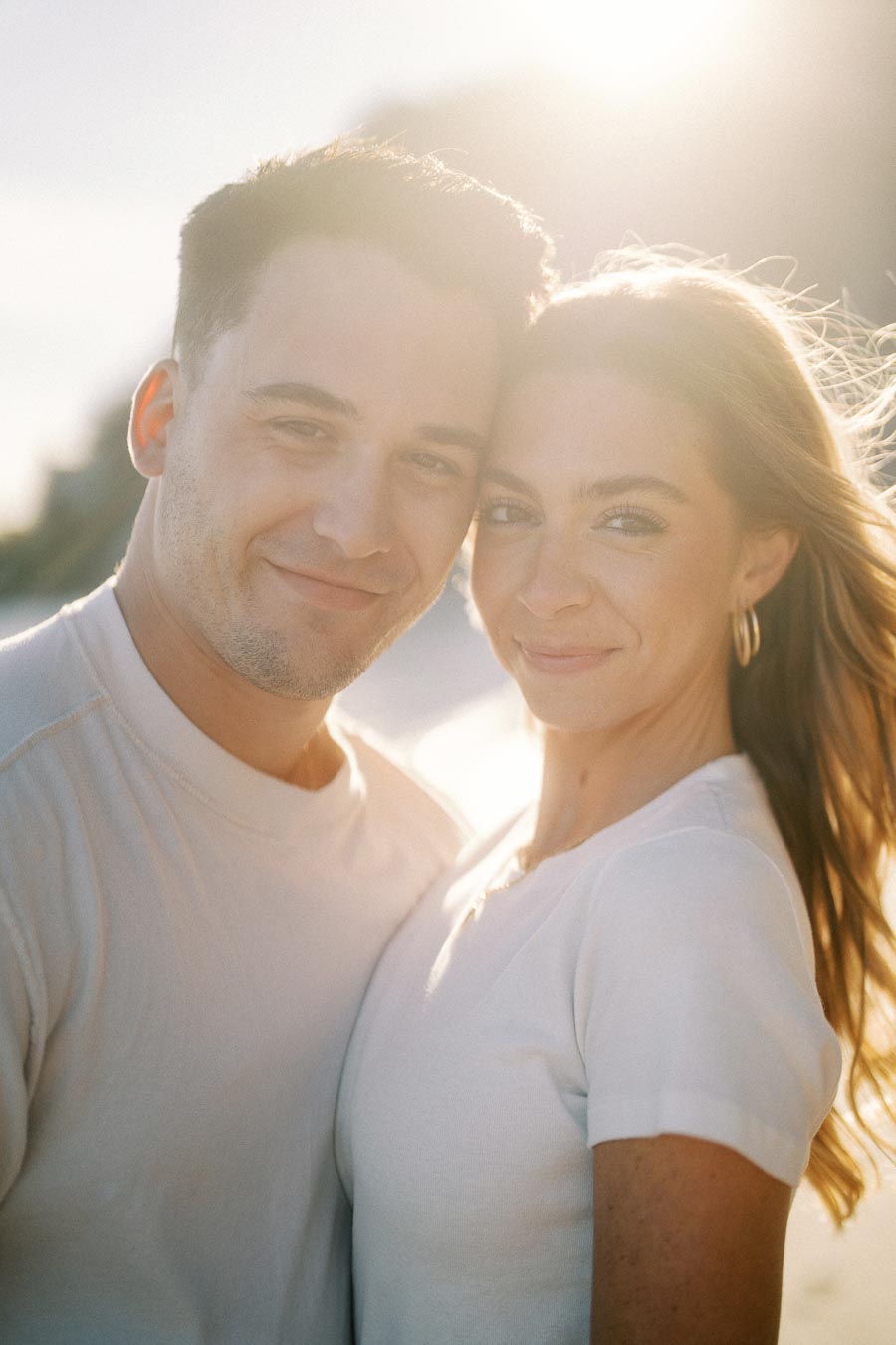A smiling couple embracing in the sunlight, both wearing white shirts, with a warm glow and soft focus background, conveying a sense of happiness and togetherness.