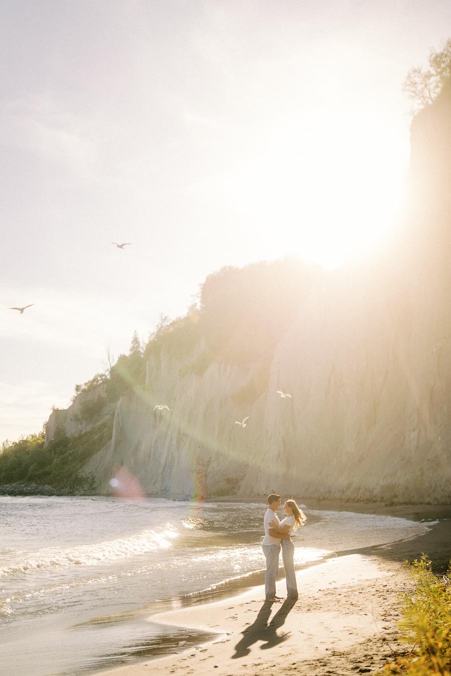 Romantic couple embracing on a sunlit beach with cliffs and seagulls in the background.