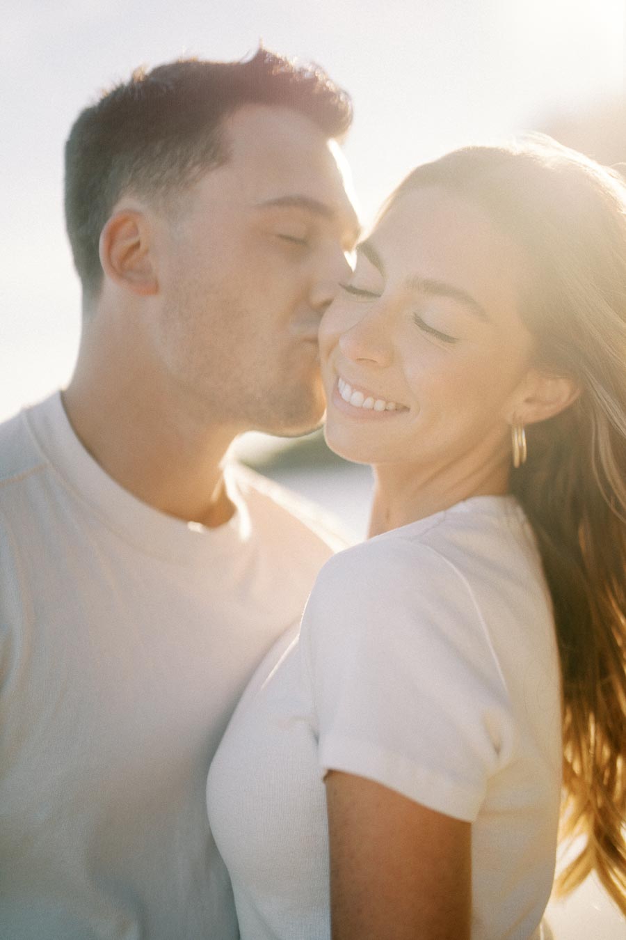 A couple sharing a tender moment as the man kisses the woman's cheek, both smiling and enjoying a sunny day outdoors, wearing casual white shirts.
