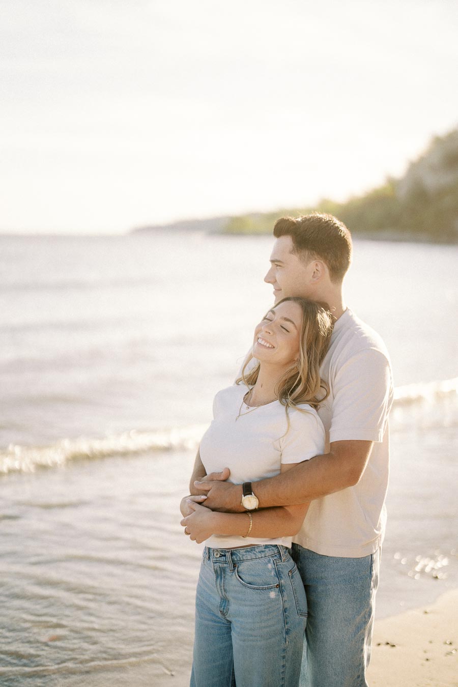 A couple embracing and smiling while standing on a sunny beach, wearing casual white shirts and jeans, with gentle ocean waves in the background.