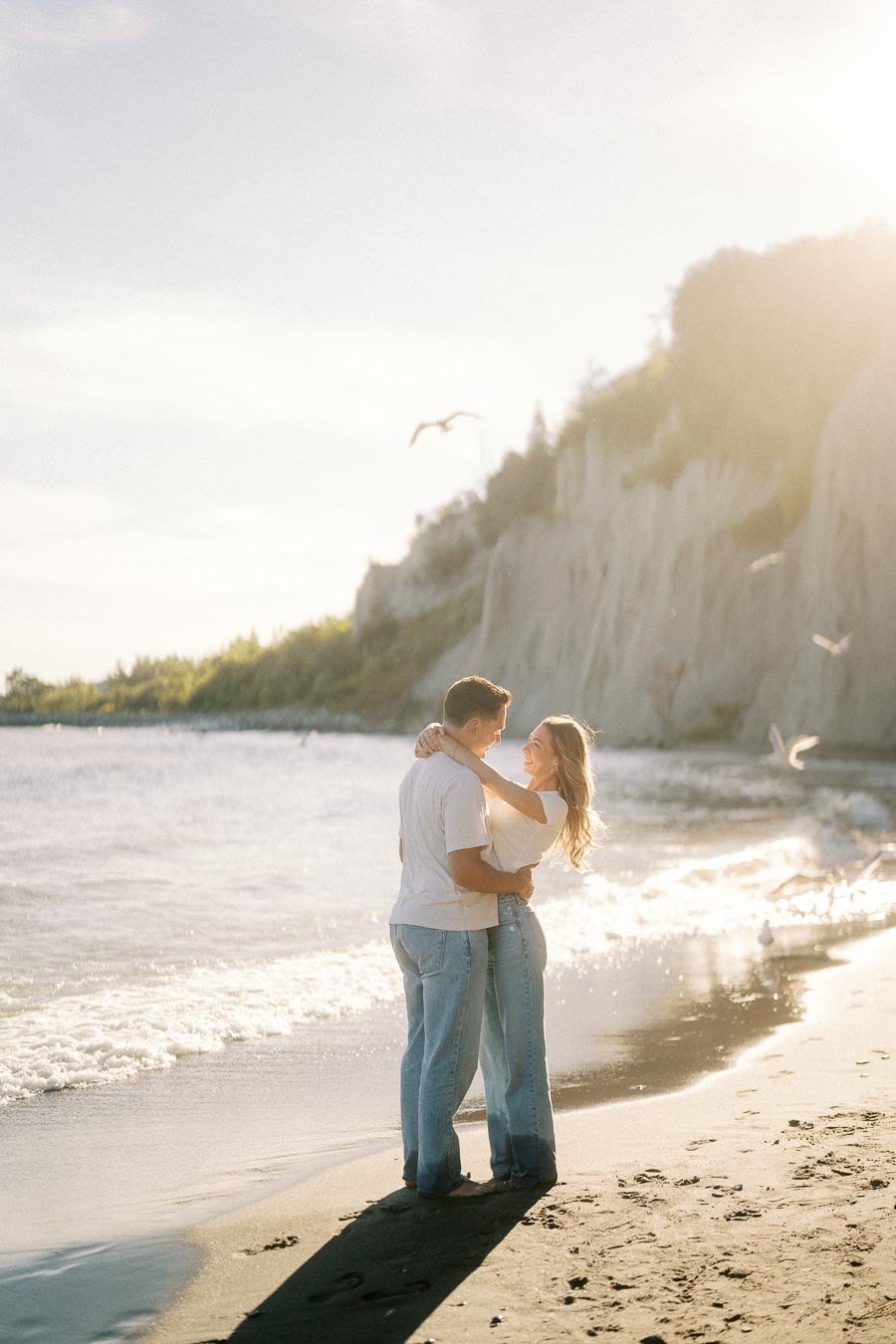 A couple embracing on a sunlit beach with waves gently crashing beside them and birds flying overhead, in front of a scenic cliffside.