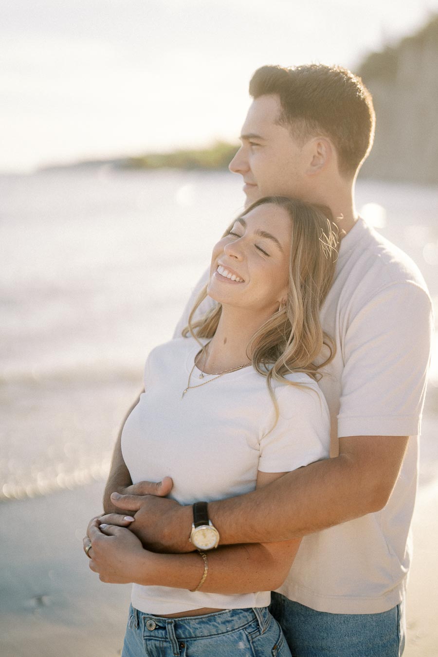 Happy couple embracing on a sunny beach, with the woman smiling and the ocean in the background.