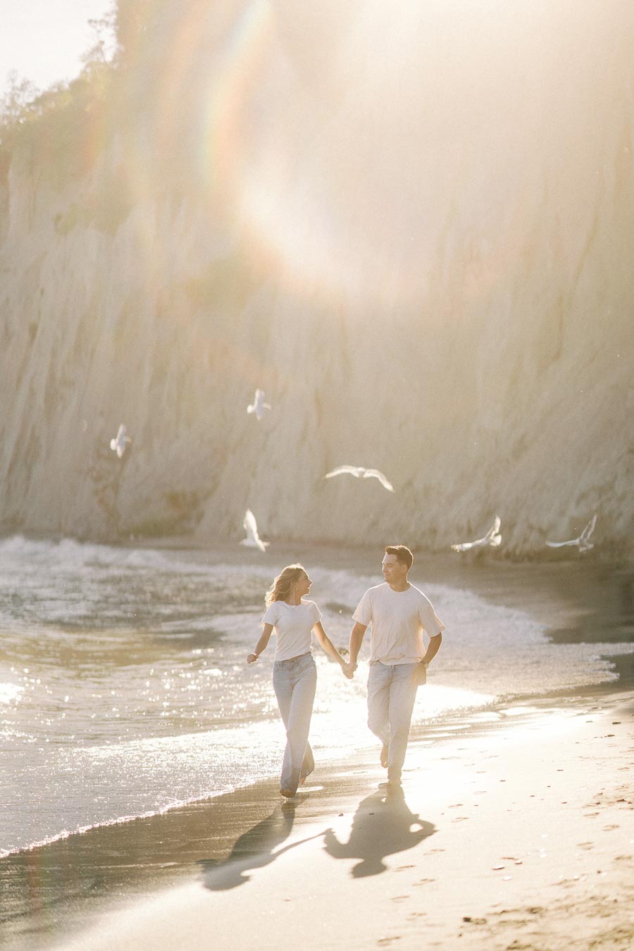 A couple holding hands and running along a sunlit beach, surrounded by seagulls, during a golden hour. Glistening waves lap at the shore with cliffs in the background, creating a serene and romantic coastal scene.