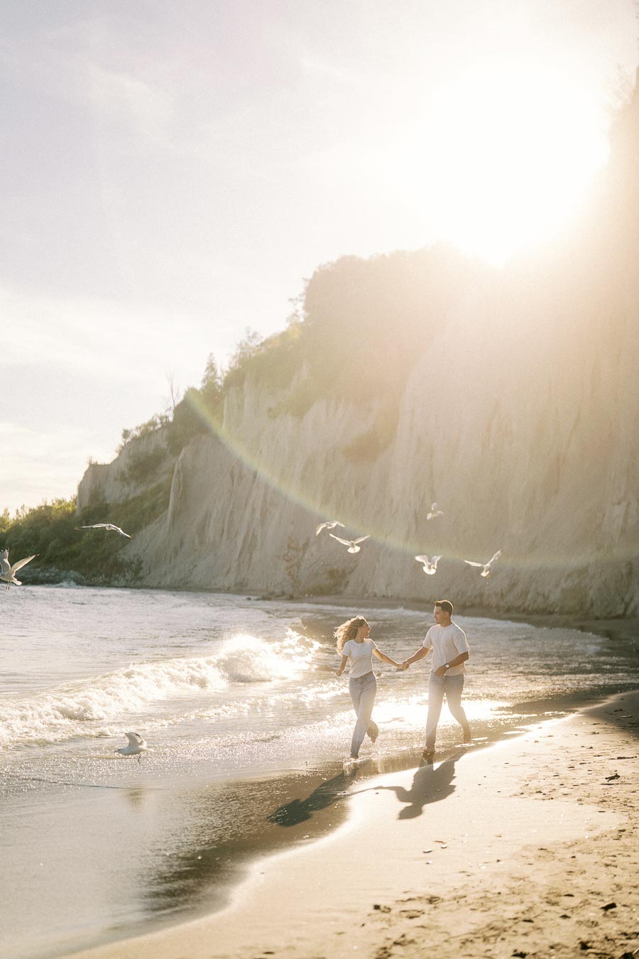 A couple holding hands and running along a sunlit beach with seagulls flying overhead and cliffs in the background.