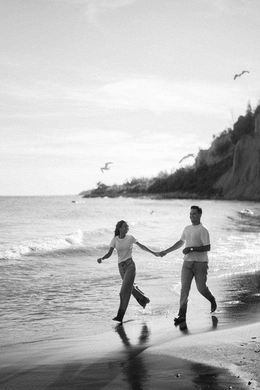 A black and white image of a couple joyfully running and holding hands along a beach shoreline, with gentle waves and birds in the background.