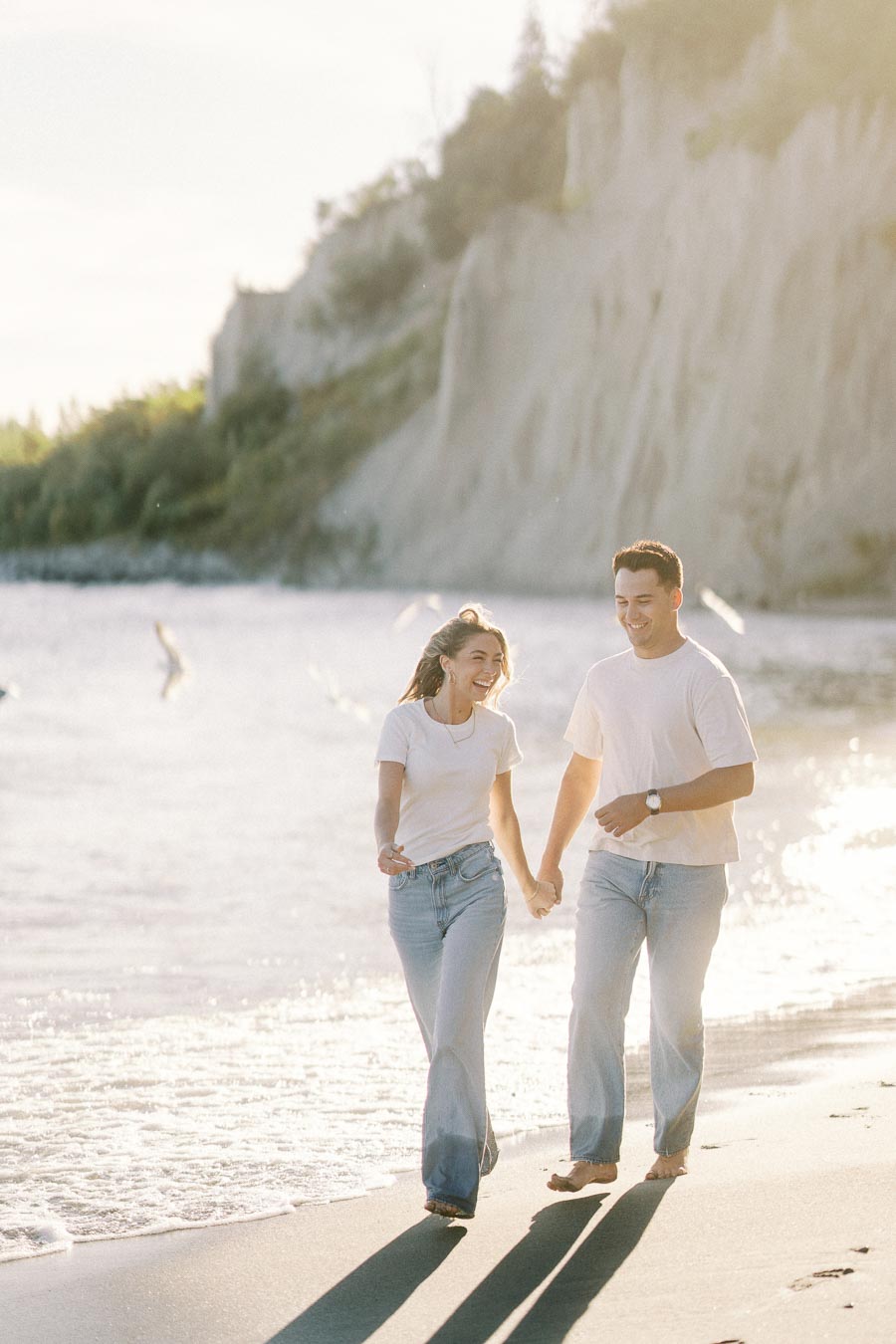 A couple holding hands and laughing while walking along a sandy beach, with cliffs and gentle waves in the background under a warm sunset glow.