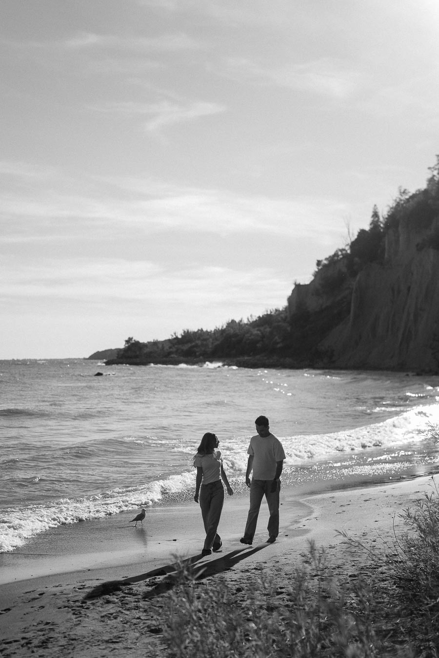 A black and white image of a couple walking along a scenic beach with gentle waves and a rocky cliffside in the background. The bright sky and ocean create a serene, romantic atmosphere.