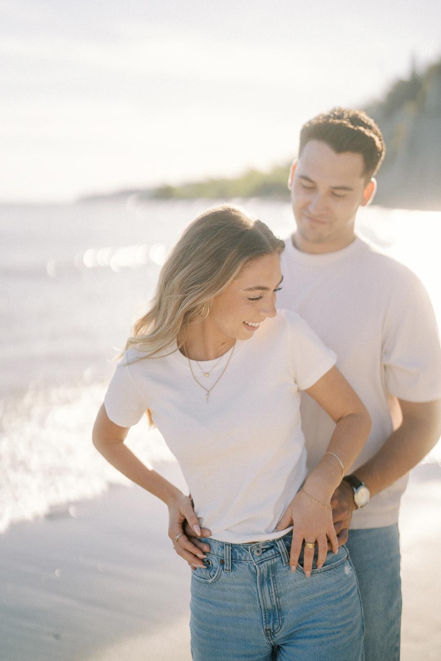 Couple embracing on a sunny beach, smiling and casually dressed in white shirts and jeans, with gentle waves in the background.