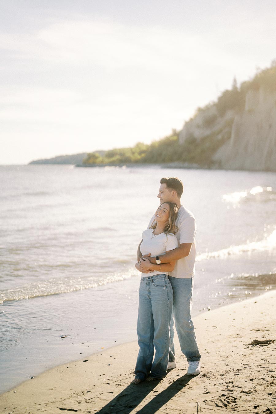 A happy couple embracing on a sunny beach with gentle waves in the background, wearing casual clothing and enjoying a peaceful moment together.