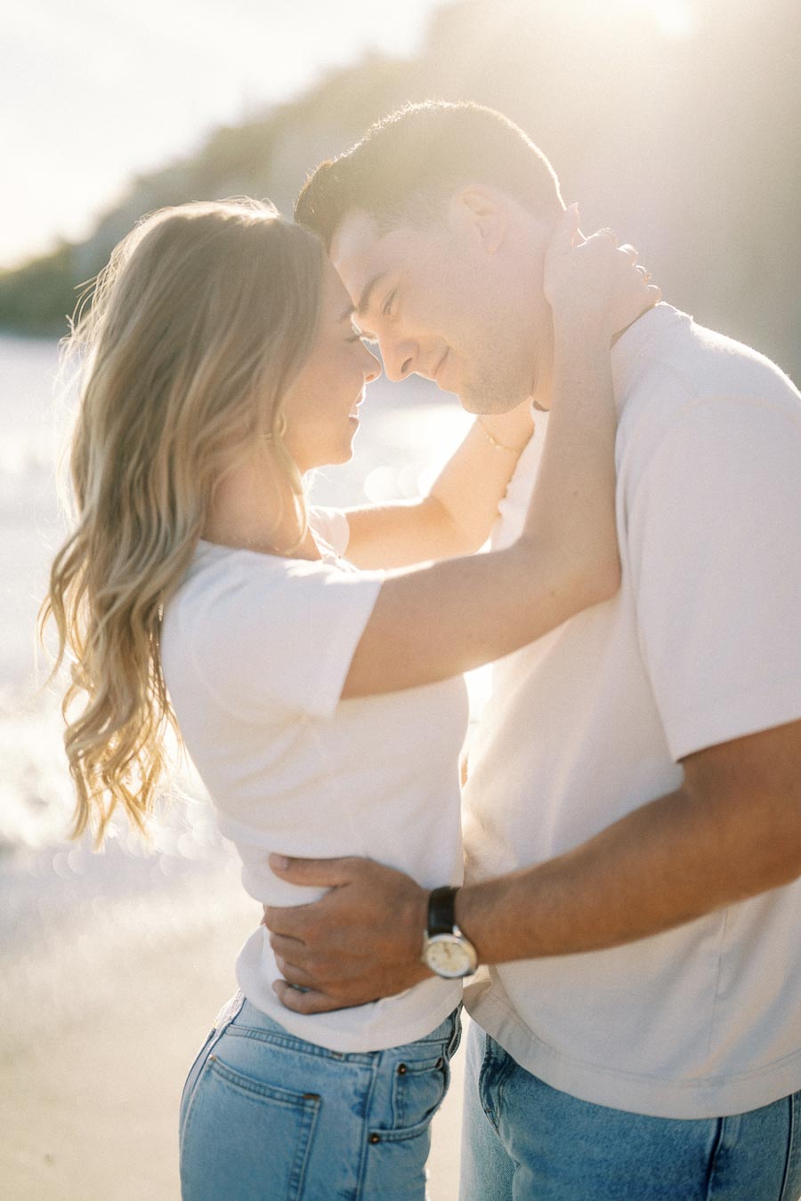 A couple embracing lovingly on a sunlit beach, both wearing casual white shirts and blue jeans, with sunlight creating a warm, romantic atmosphere.