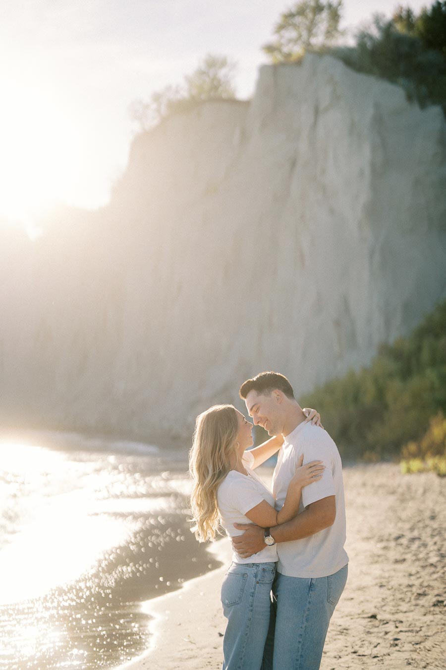 A couple embracing on a sunny beach with cliffs in the background, bathed in soft, golden sunlight.