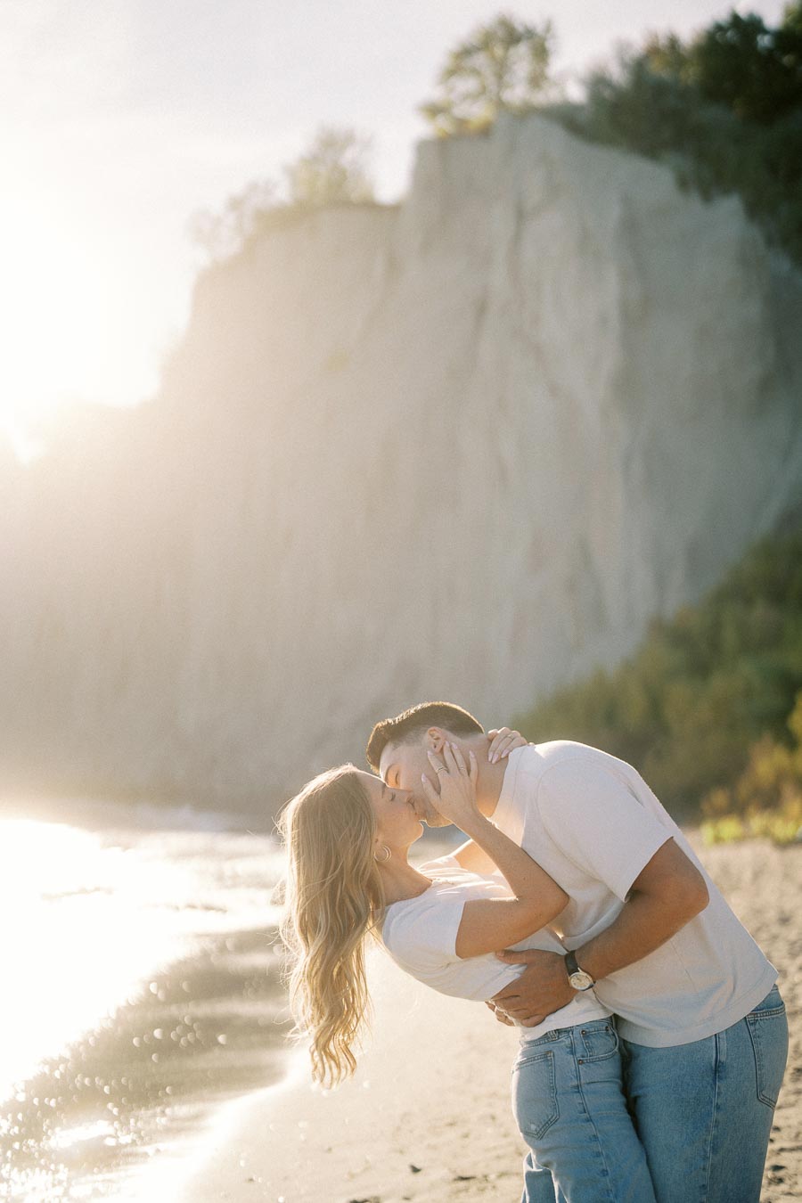 A couple sharing a romantic kiss on a sunny beach, with a cliff and clear blue sky in the background. Both are wearing casual white and blue attire, enjoying a serene, picturesque moment by the water's edge.