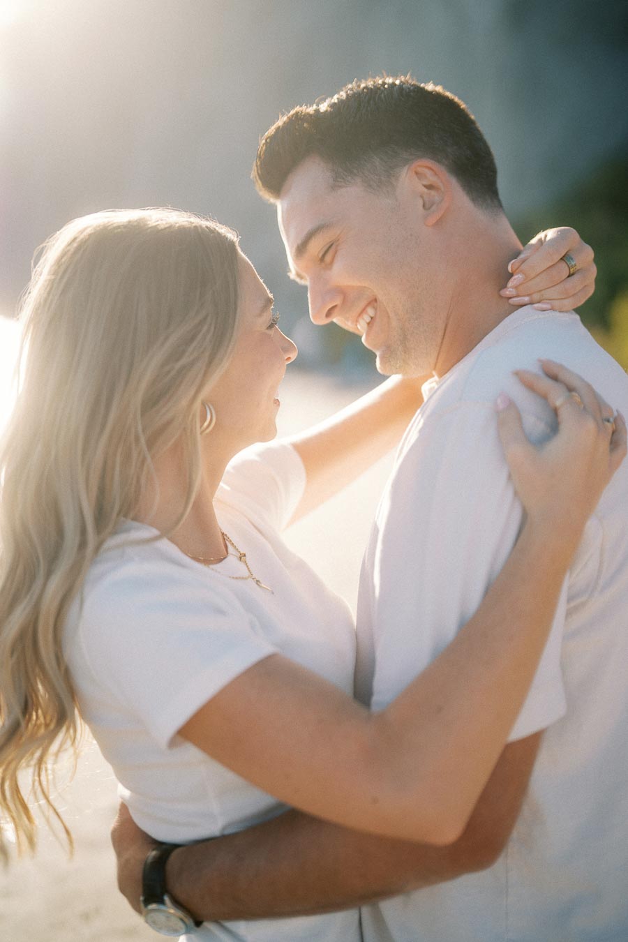 A couple embraces and smiles at each other under soft, natural sunlight outdoors.
