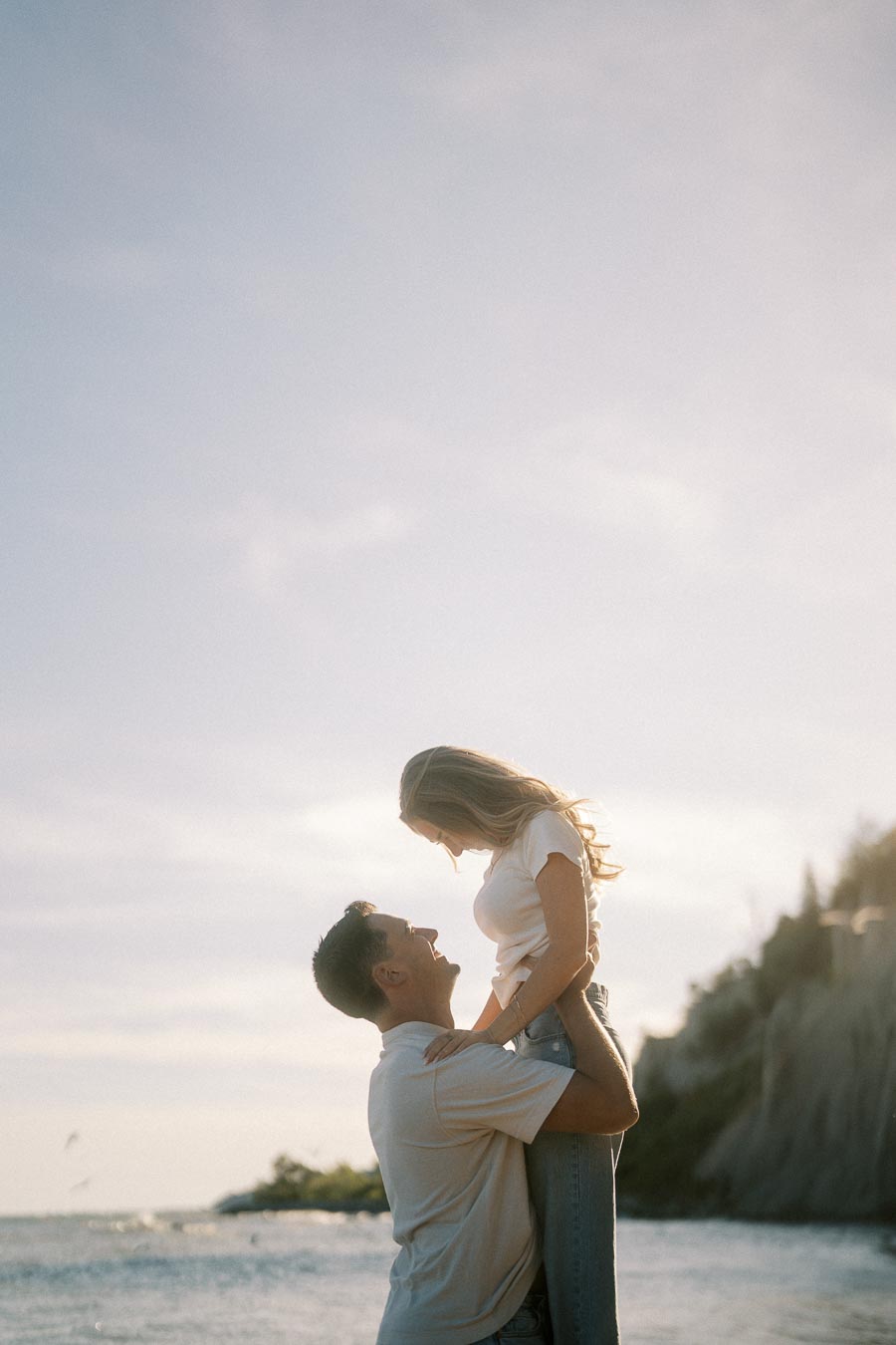 Couple enjoying a romantic moment by the seaside at sunset, with a man lifting a woman surrounded by nature and calm waters in the background.