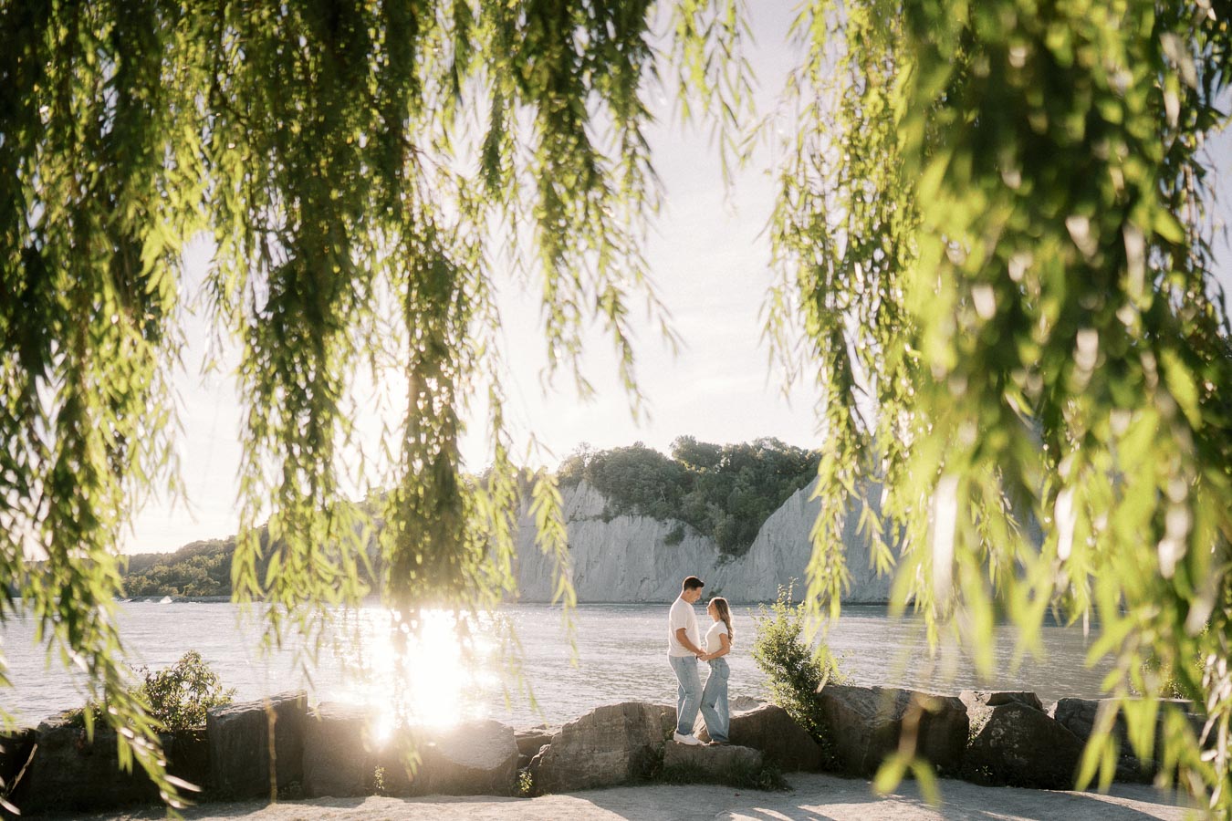 A couple holding hands by a lake with sunlight reflecting off the water, framed by lush green willow branches.