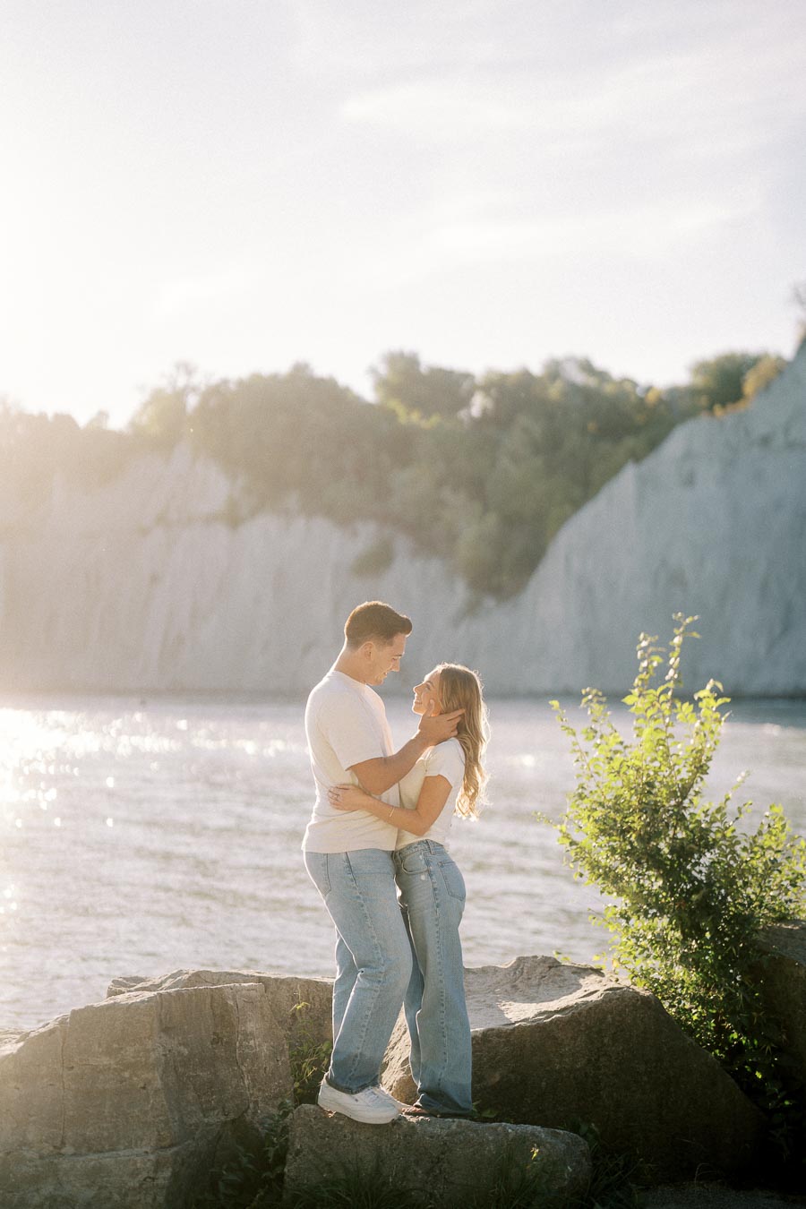 A couple embracing on a sunny day by the lake with cliffs and trees in the background.