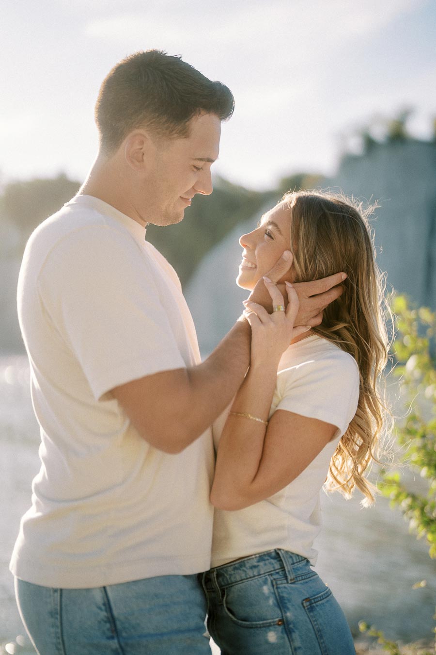 Happy couple embracing by a serene lakeside, dressed in casual white tops and jeans, capturing a romantic moment with soft natural lighting.