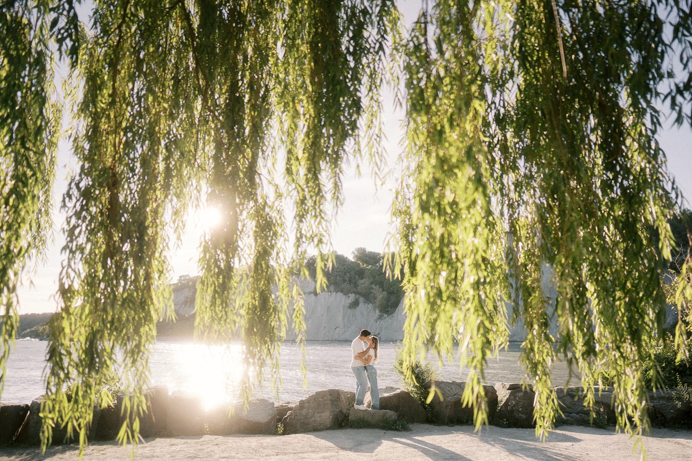 Romantic couple embraces under lush willow tree by sunlit lakeside with scenic cliffs in the background
