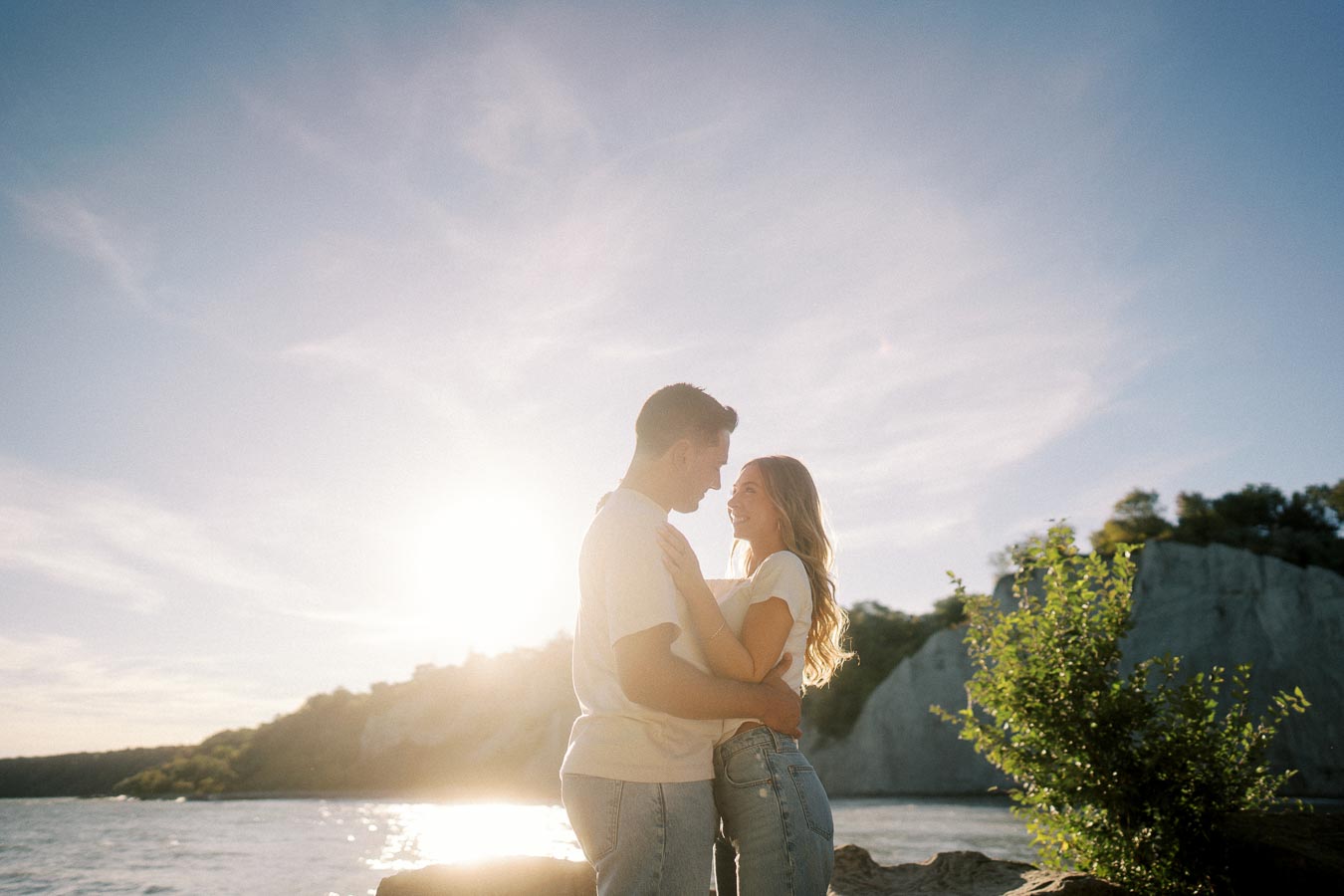 A couple embracing and smiling at each other by a scenic lakeside during sunset, surrounded by clear skies and nature.