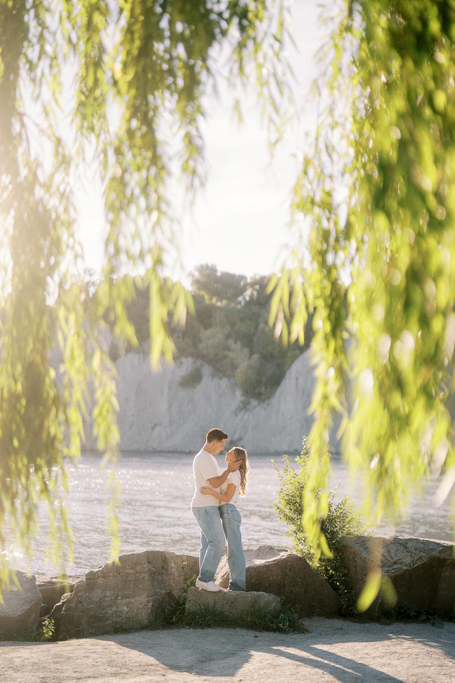 A couple embracing by a serene lakeside under lush greenery, with sunlight filtering through the leaves, creating a romantic and tranquil atmosphere.
