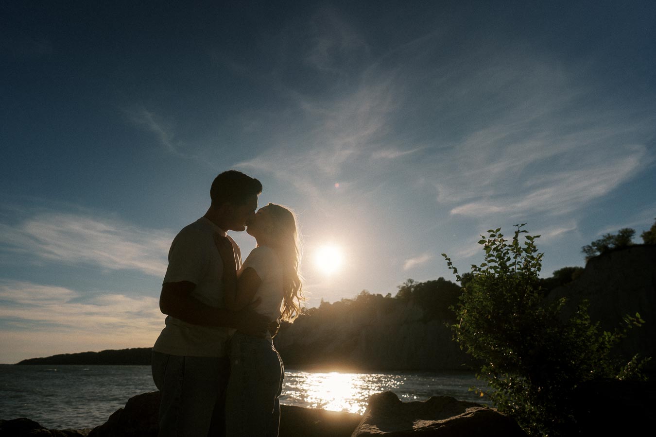 Silhouette of a couple kissing at sunset by a serene lakeside, with a clear sky and gentle waves in the background.