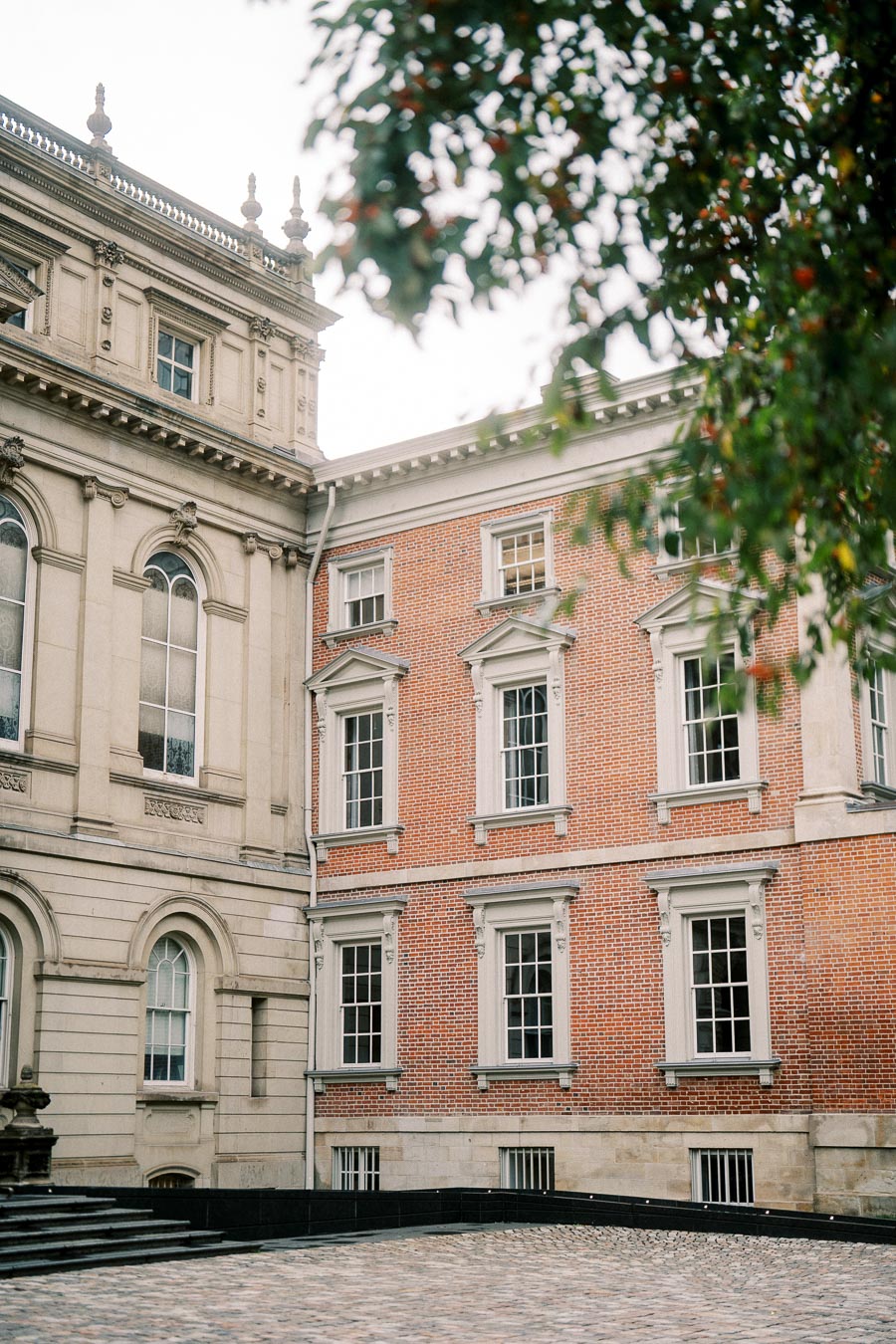 Exterior view of a historic building with ornate architecture, featuring brick and stone facades, arched windows, and a cobblestone courtyard with lush green foliage in the foreground.
