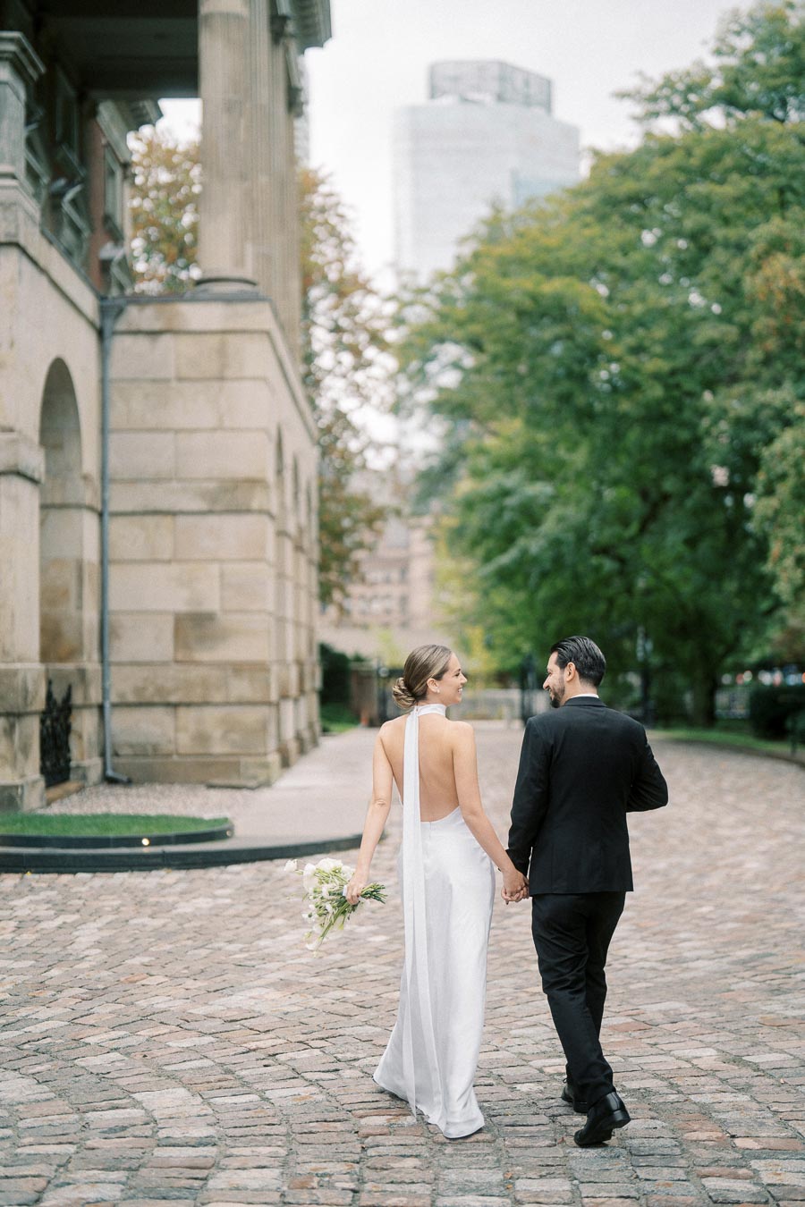 A newlywed couple walking hand in hand on a cobblestone path, with the bride in an elegant white dress holding a bouquet, and the groom in a black suit, surrounded by historic architecture and lush greenery.