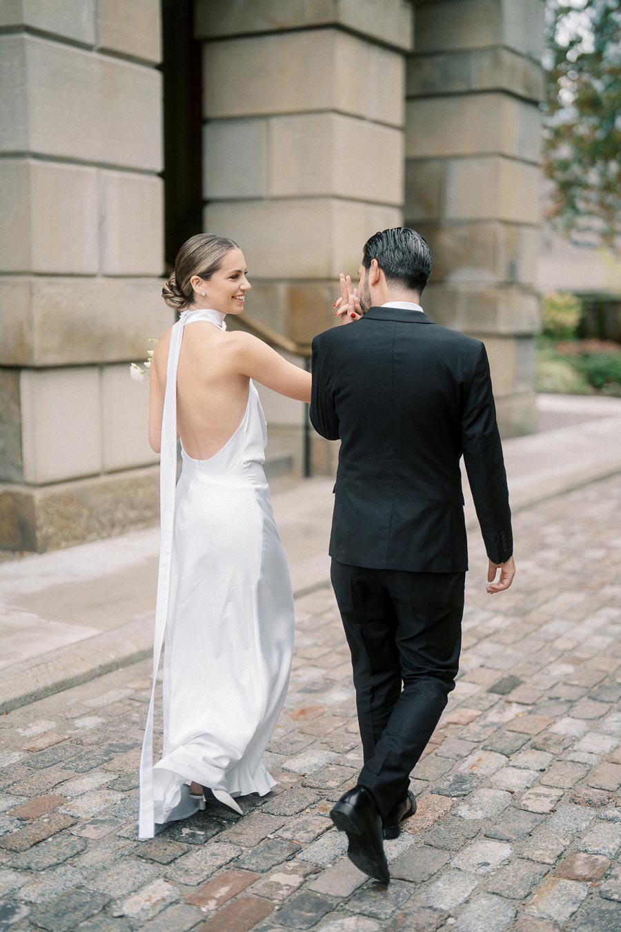 Bride and groom walking hand in hand on cobblestone street, she in a white backless dress and he in a black suit, with elegant architectural backdrop.