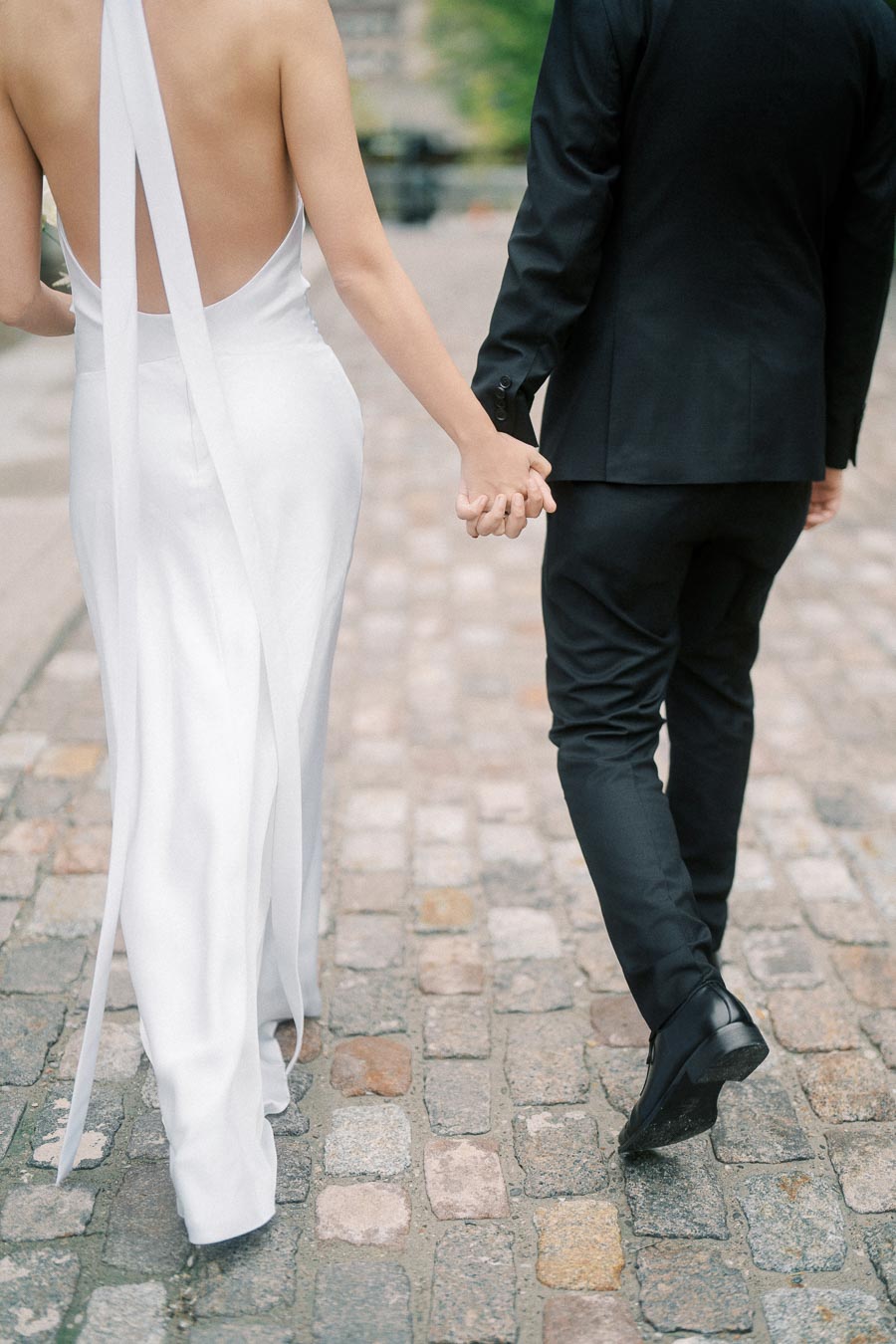 Rear view of a newlywed couple in formal attire holding hands on a cobblestone street.
