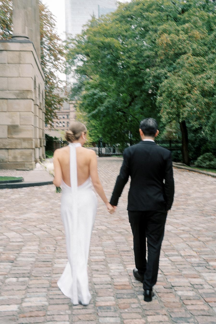 A bride in an elegant white gown and a groom in a black suit hold hands while walking on a cobblestone path surrounded by greenery and historic architecture.