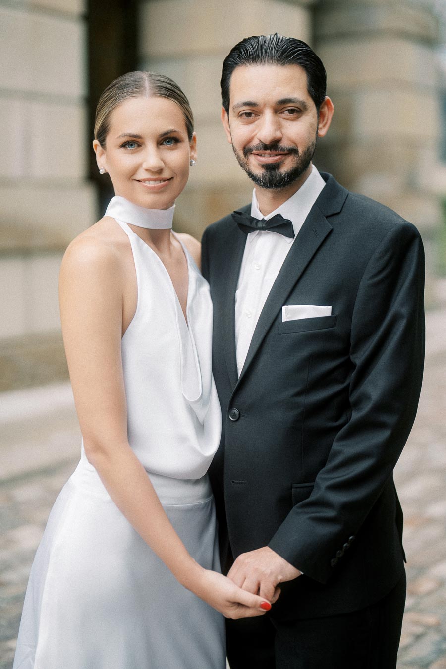 A smiling couple in formal attire, with the woman in a white dress and the man in a black suit and bow tie, holding hands outdoors.