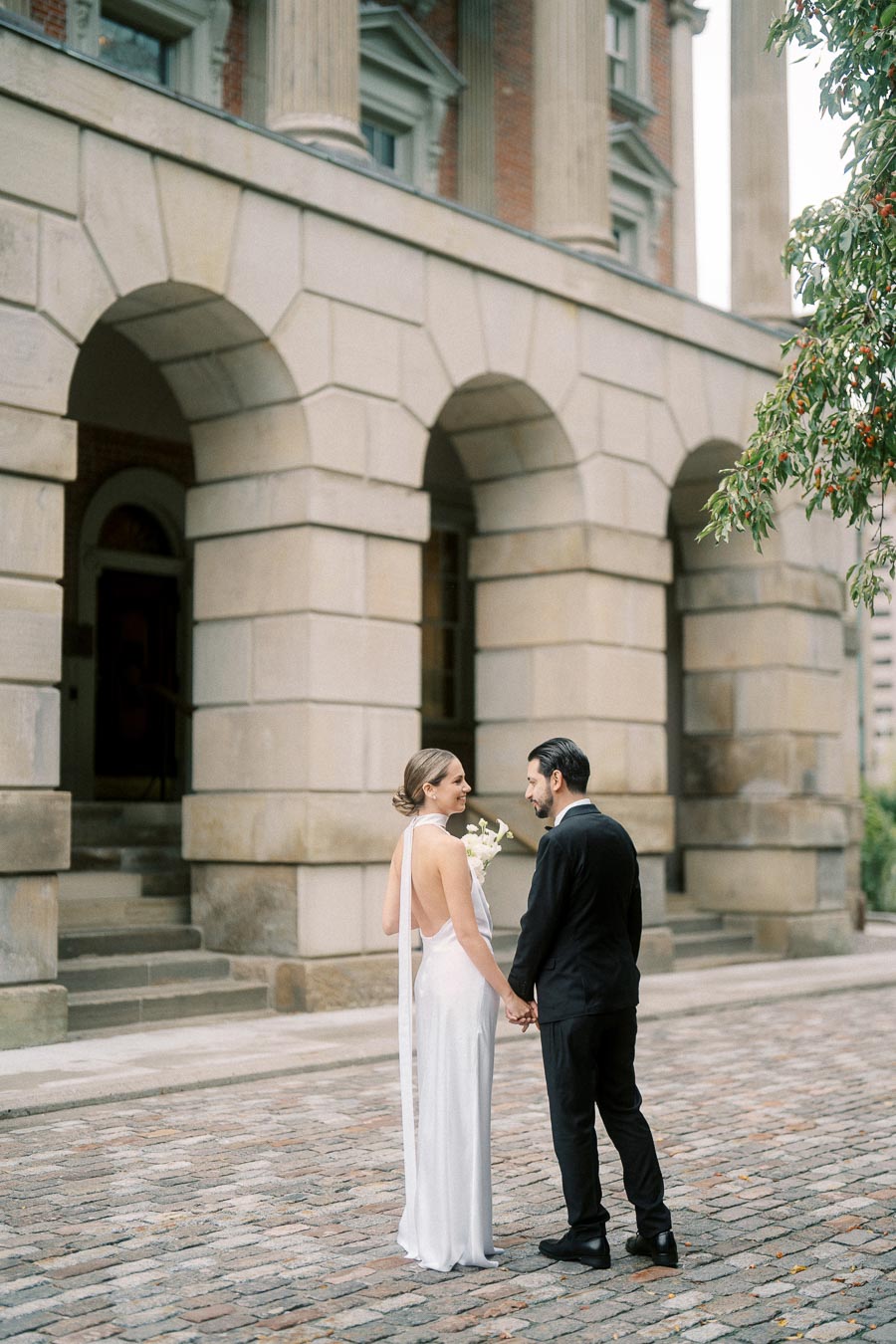 Elegant couple in wedding attire holding hands outside a historic building with arched entrances and cobblestone path, capturing a romantic moment.