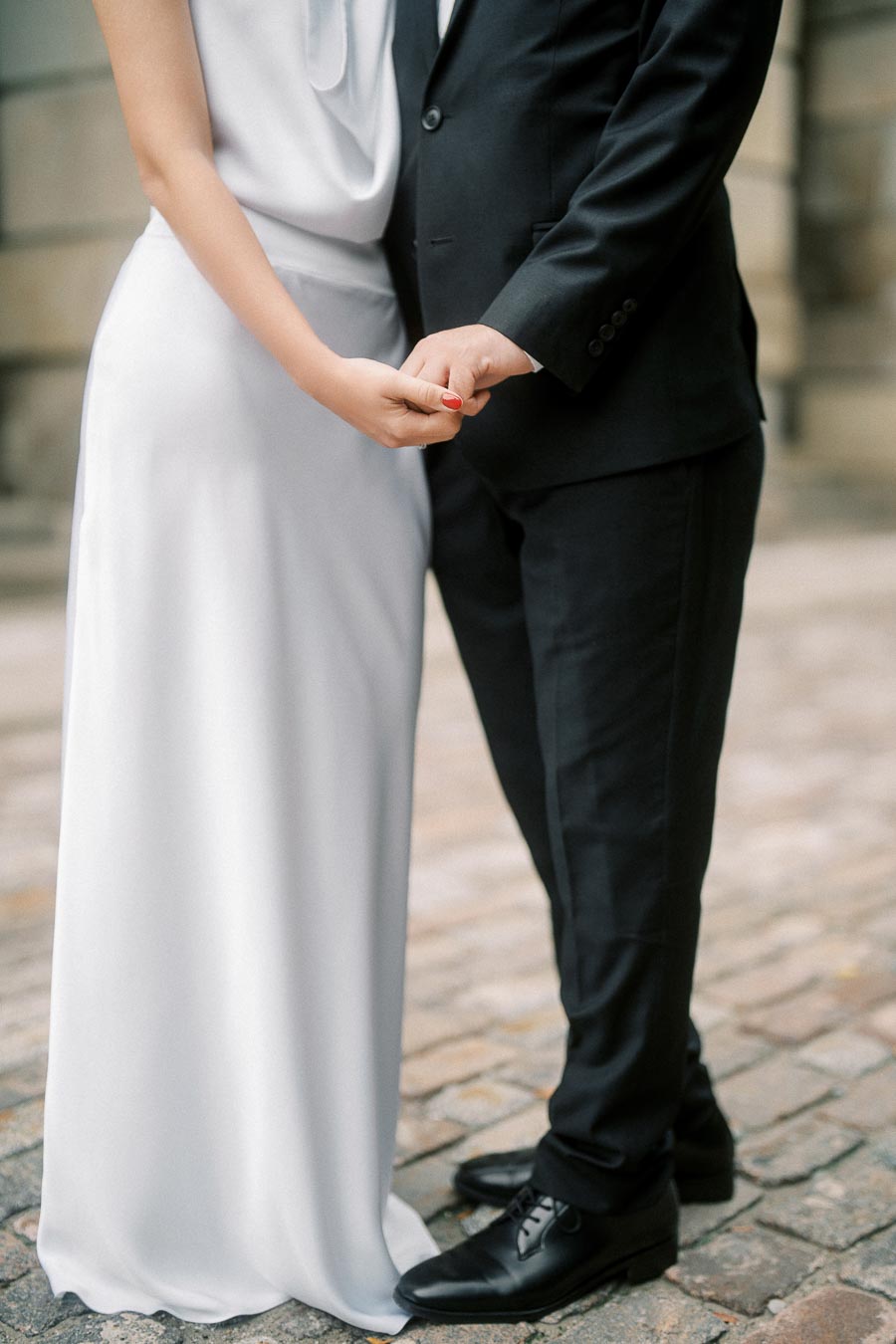 A couple holding hands in elegant wedding attire, standing on cobblestone pavement.