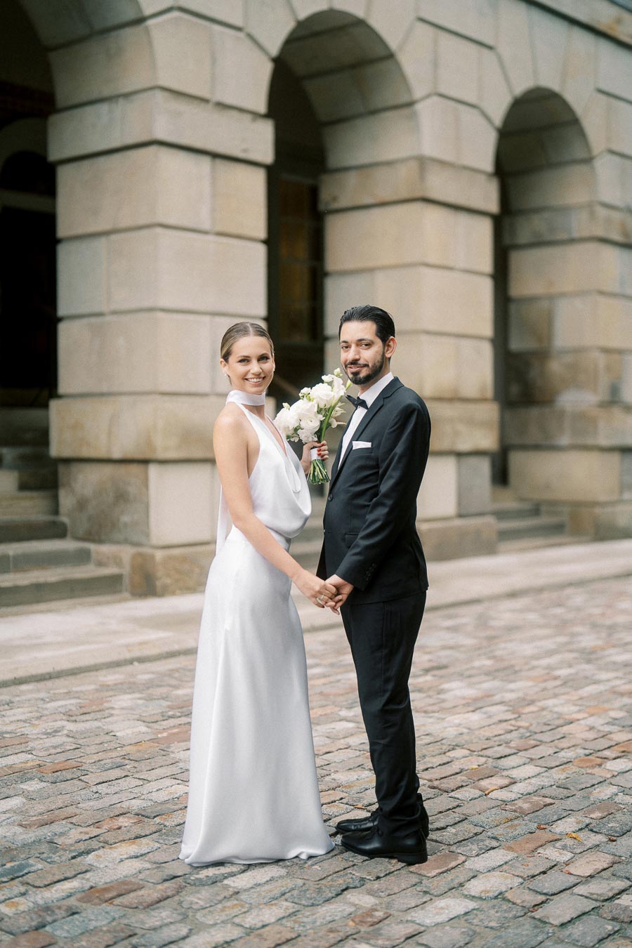 Elegant couple in wedding attire holding hands, standing on cobblestone pavement in front of an archway.