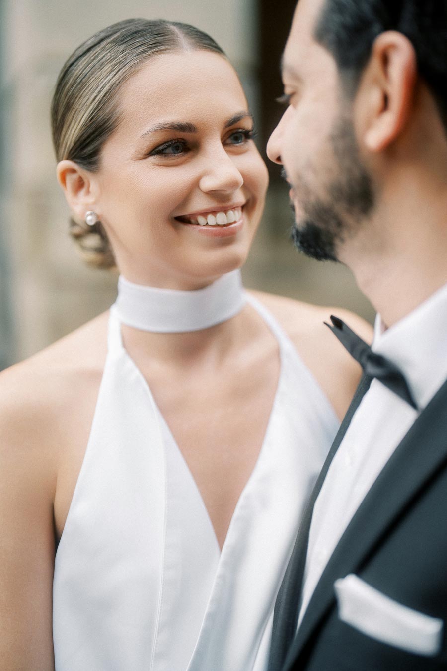 A joyful bride in a white wedding dress shares a loving gaze with the groom, dressed in a black tuxedo, highlighting a romantic wedding moment.