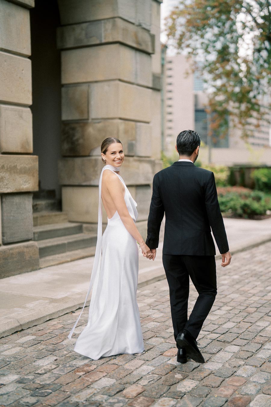 Elegant bride in white gown holding hands with groom in black suit on a cobblestone path, urban wedding setting.