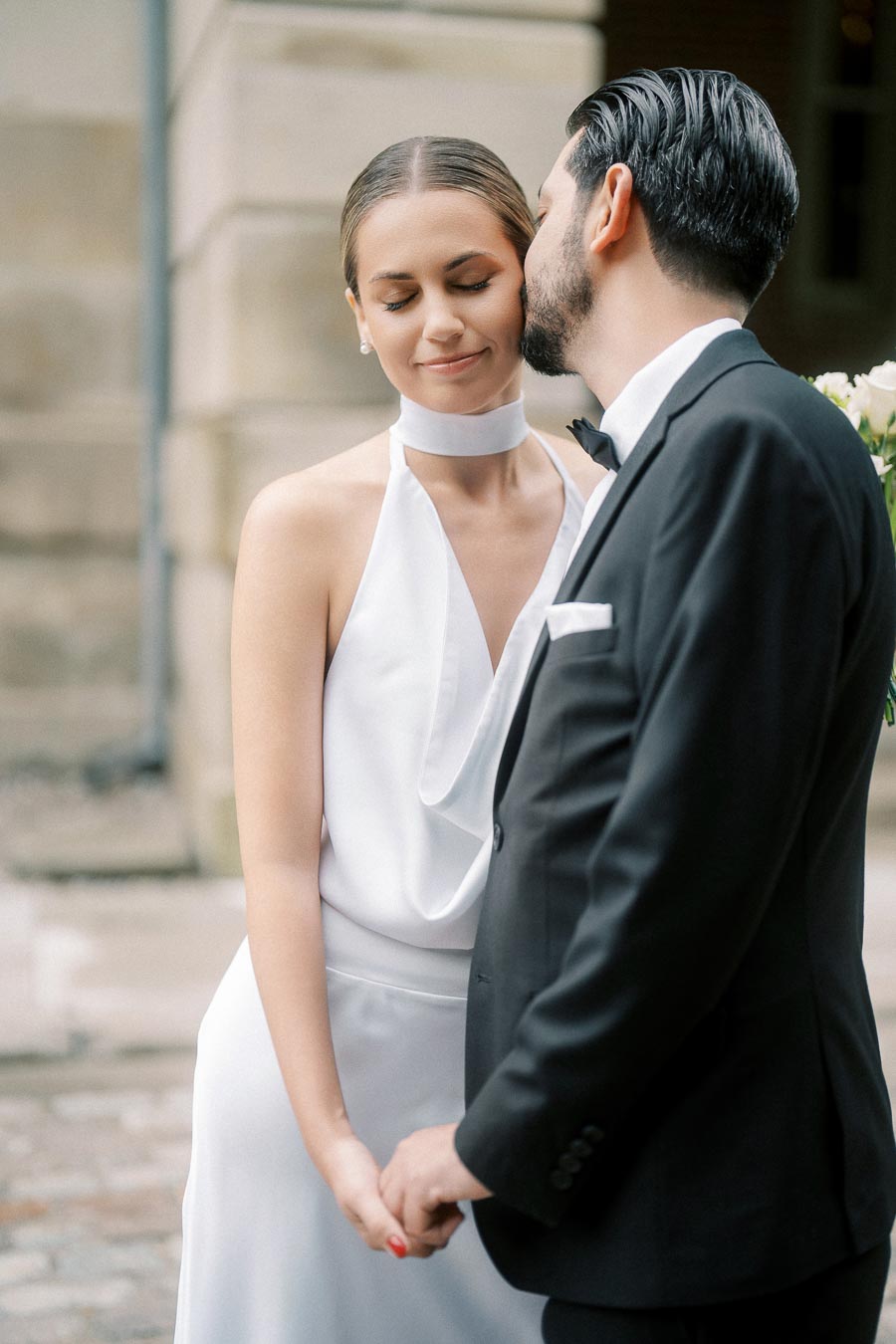 Bride in elegant white gown standing lovingly with groom in black tuxedo, sharing a tender moment during their wedding.