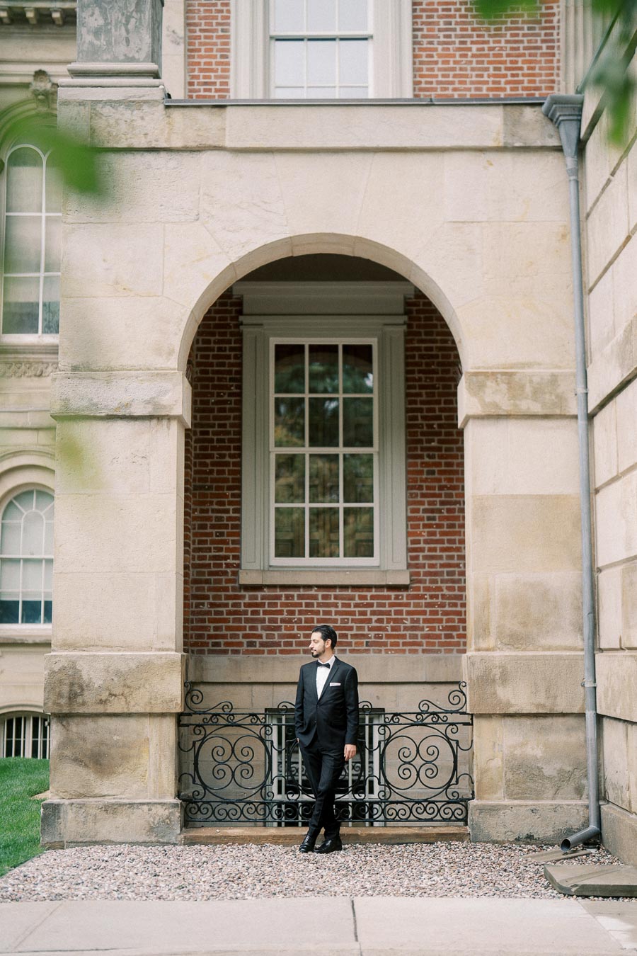 Man in formal suit stands confidently under an elegant stone archway, flanked by a red brick wall, conveying sophistication and charm in a historic setting.