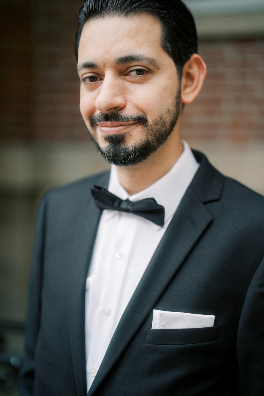 A man in a black tuxedo with a bow tie and pocket square smiles confidently, standing against a blurred brick wall background.