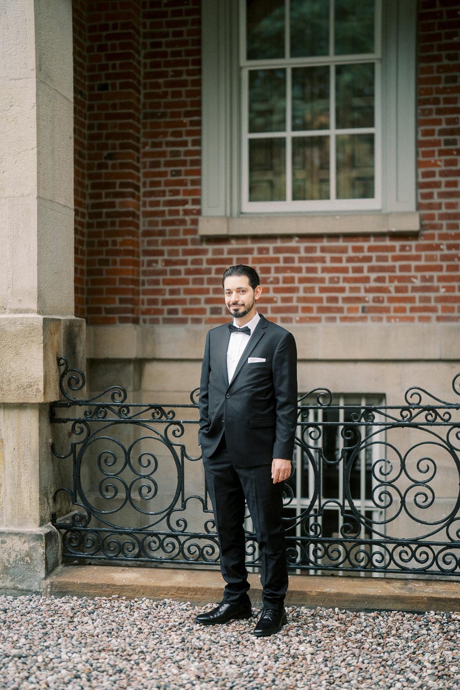 A man in a stylish black tuxedo stands confidently on a gravel pathway in front of an elegant brick wall and ornate iron railing, exuding sophistication and charm.