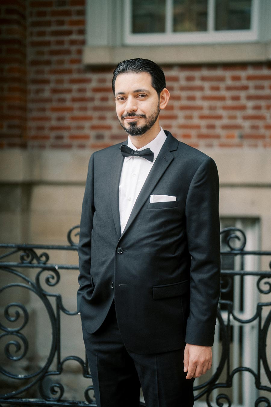 A man in a black tuxedo with a bow tie stands outdoors in front of a decorative wrought iron railing and brick wall, smiling confidently.