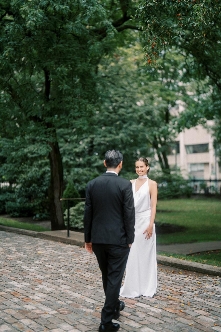 A bride in a white dress smiles at a groom in a black suit as they stand on a cobblestone path surrounded by lush green trees.