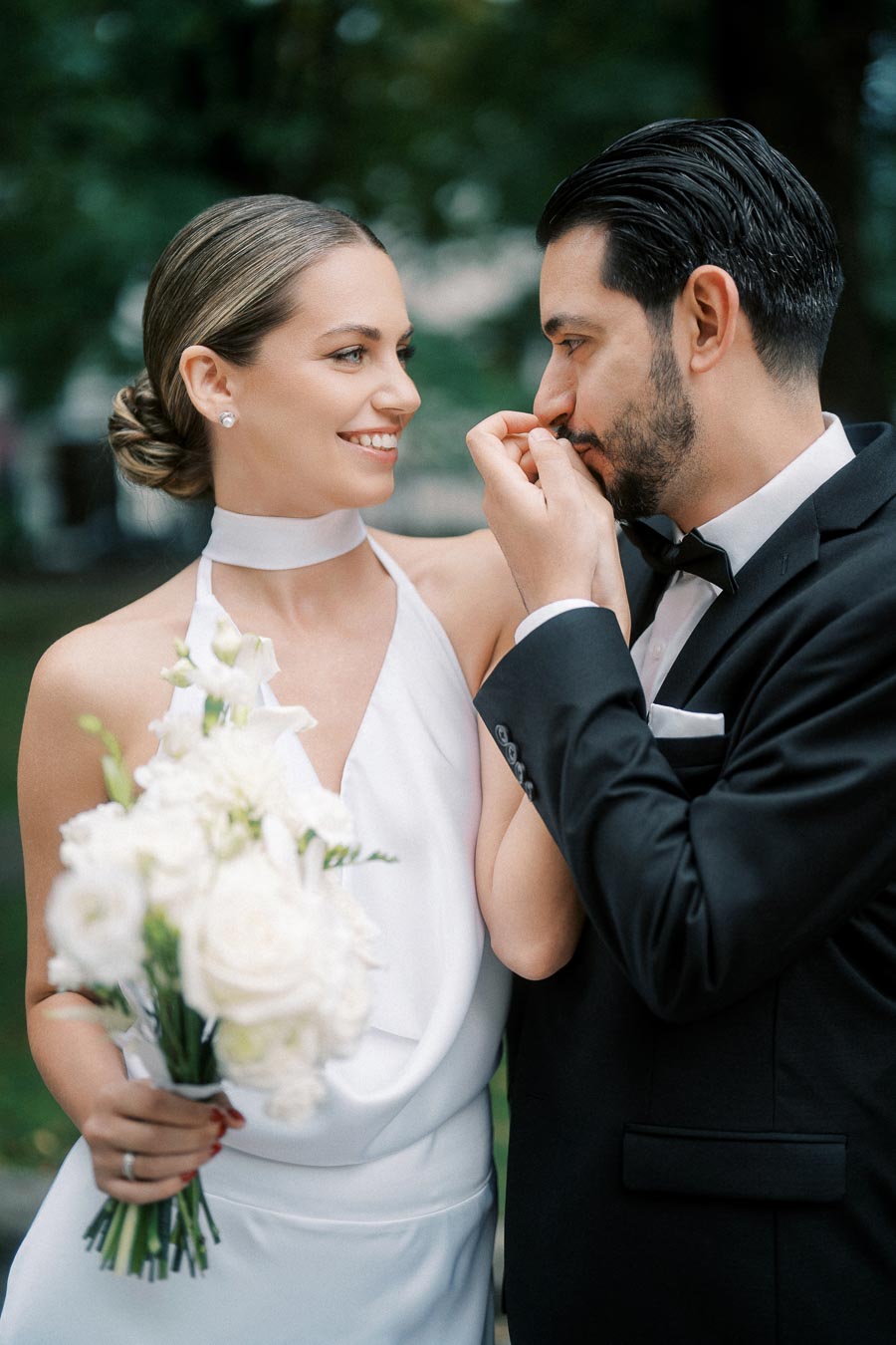 A joyful couple on their wedding day, with the bride holding a bouquet of white flowers and the groom gently kissing her hand. The bride is in a white dress and the groom in a black tuxedo, set against a blurred natural background.