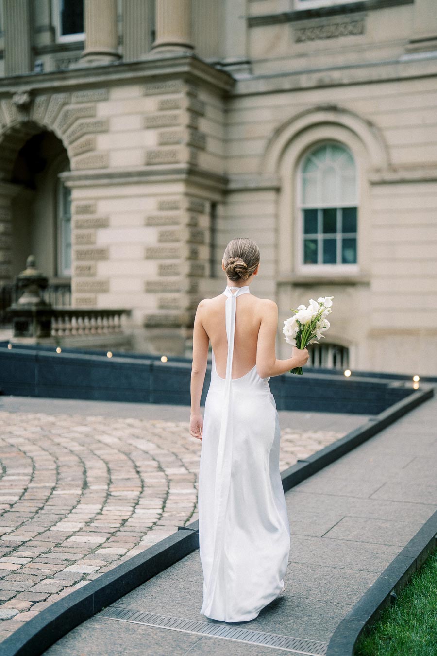 A bride in an elegant, backless white wedding dress walks along a cobblestone path with a bouquet of white flowers, set against a historic stone building backdrop.
