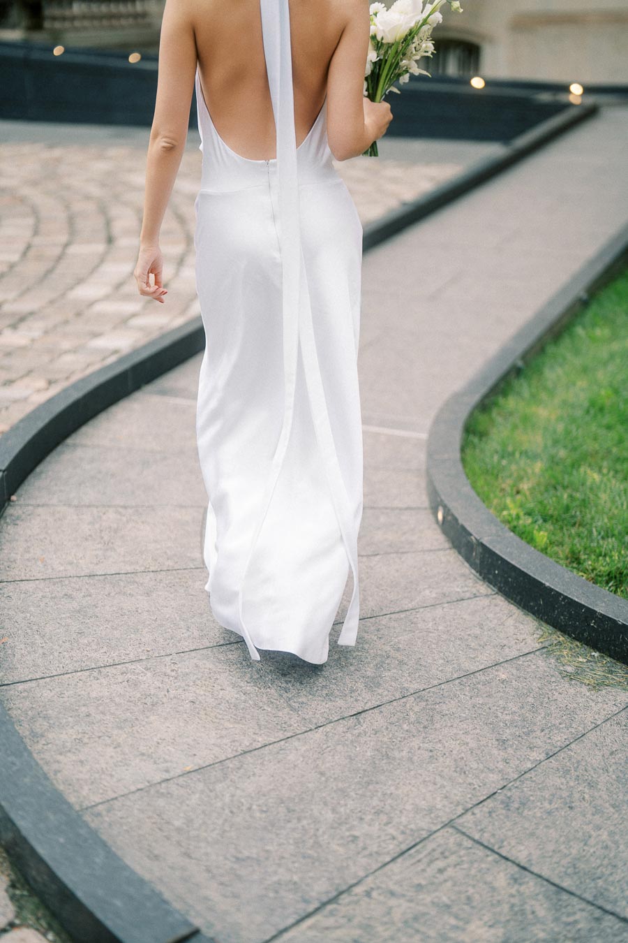 Bride in elegant backless white wedding gown walking down a curved stone pathway, holding a bouquet of white flowers.