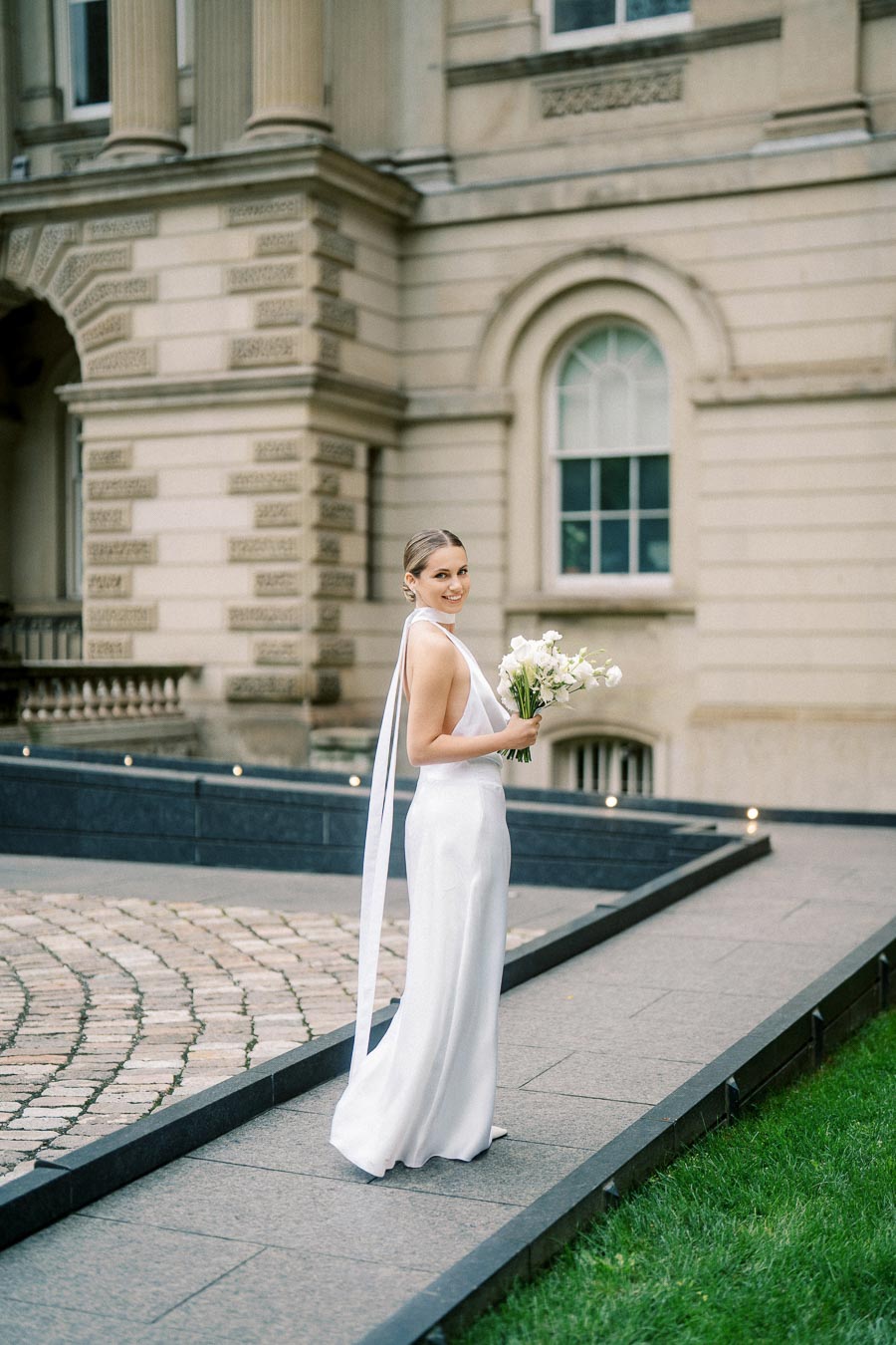 Bride in elegant white gown holding bouquet, posing outside historical building with intricate architecture, showcasing a classic wedding scene.