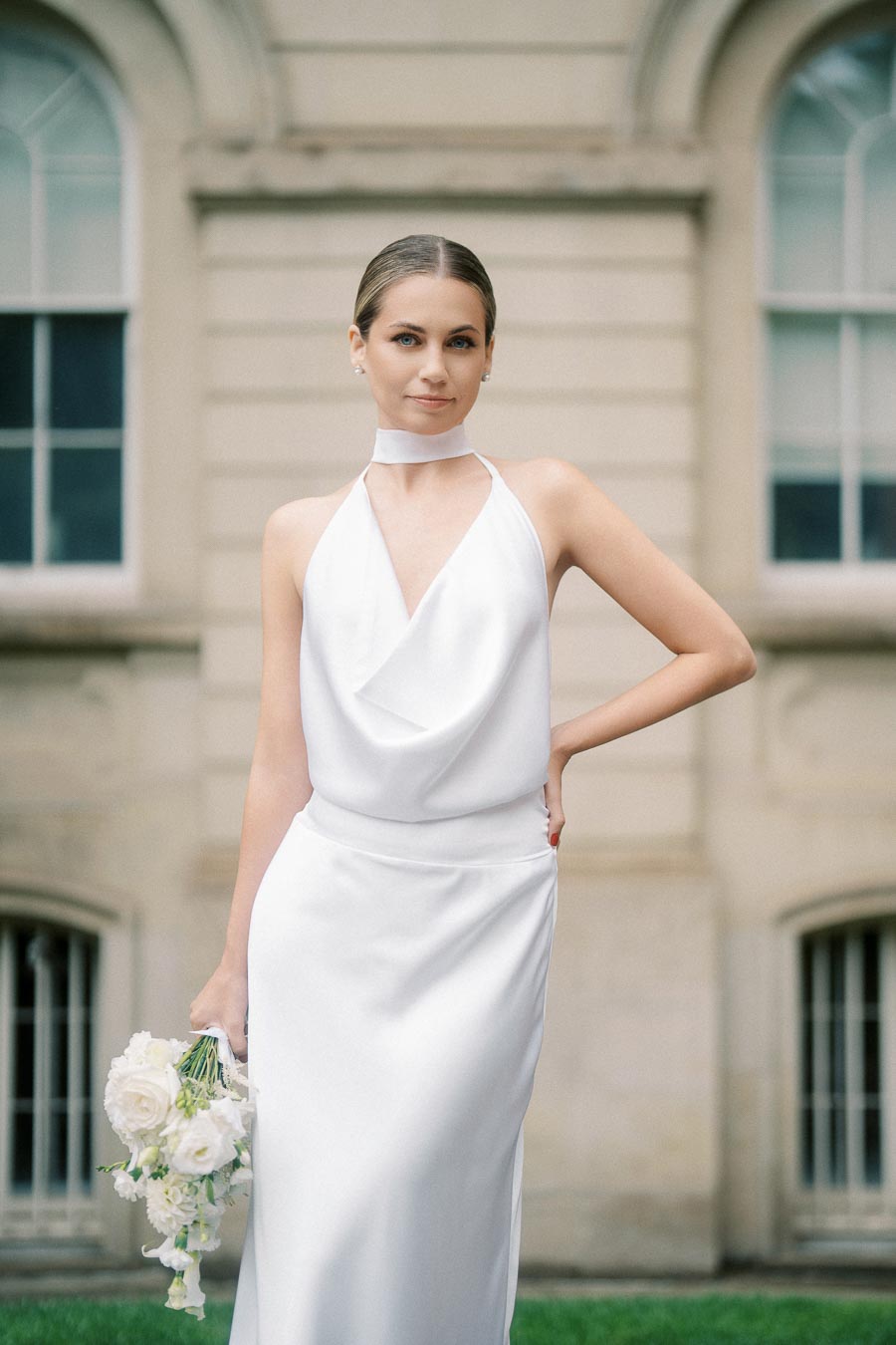 A bride in a stylish halter-neck white wedding dress holding a bouquet of white flowers, standing in front of a historic building with arched windows.