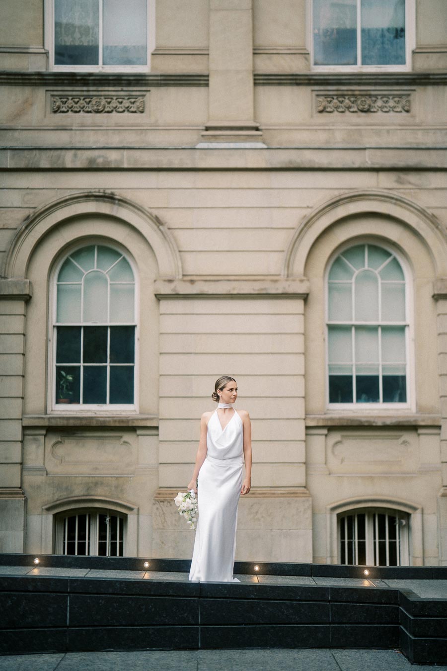 Elegant bride in a white gown holding a bouquet, standing in front of a historic building with arched windows.