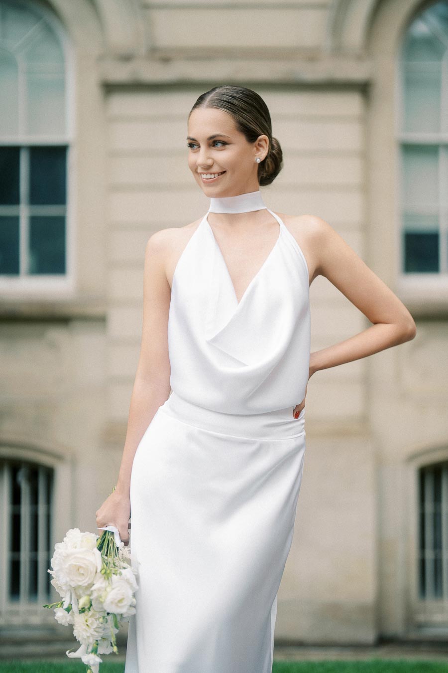 Bride in elegant white halter-neck wedding dress holding a bouquet of white flowers, posing outdoors in front of a historic building.