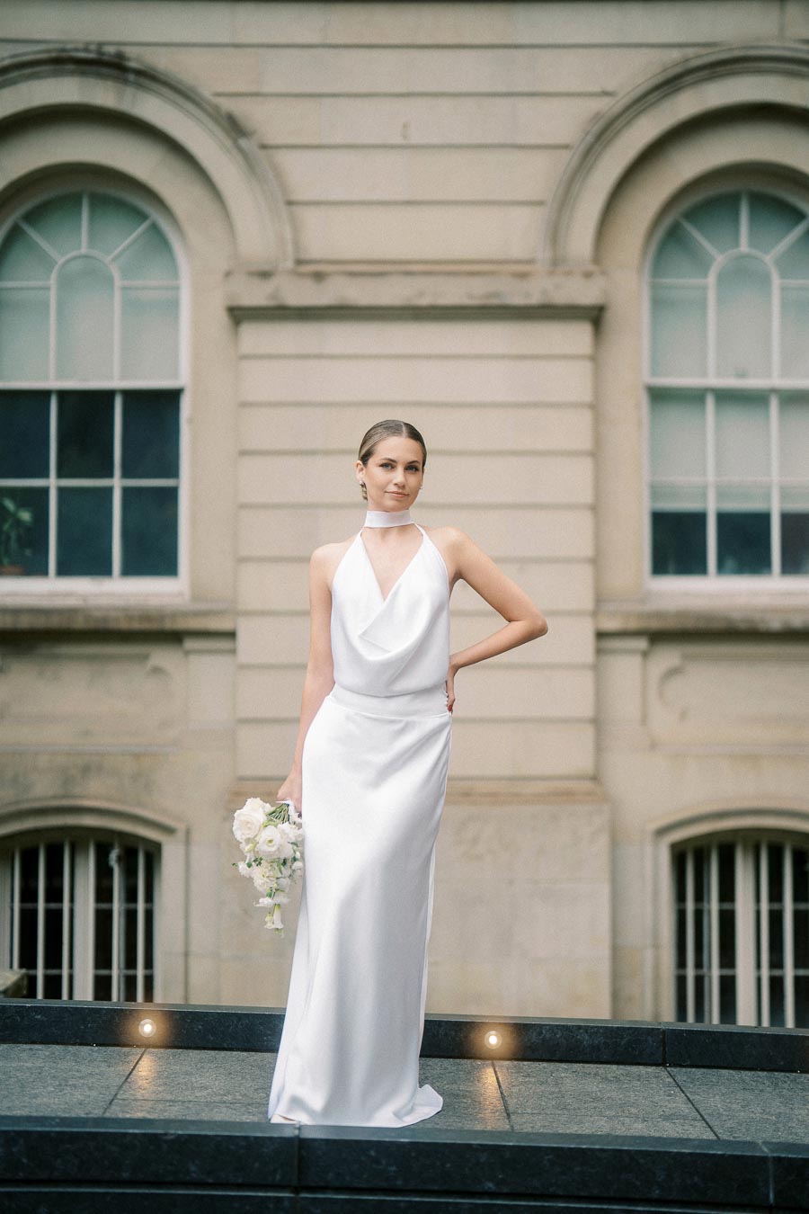 A bride in an elegant white halter-neck wedding dress holding a bouquet of white flowers stands confidently outdoors in front of an architectural building with arched windows.